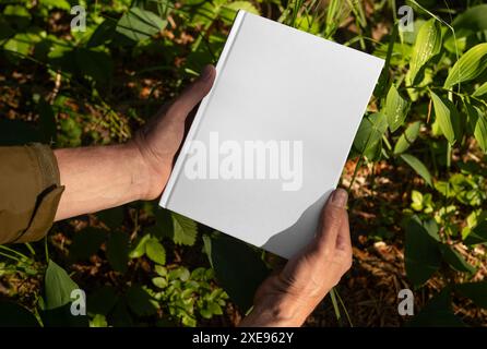 . Mani maschili che tengono il libro mockup con copertina rigida, mock-up con copertina rigida vuota in natura, piante verdi. Foto Stock
