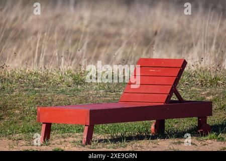 Un lettino da sole in legno rosso sdraiato si erge sull'erba in una giornata estiva di sole Foto Stock