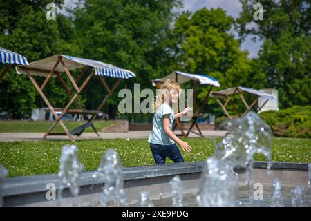 una giovane ragazza con i jeans e una camicia azzurra fa scorrere le dita attraverso l'acqua della fontana, il suo sorriso radioso sotto il sole di mezzogiorno. Foto Stock