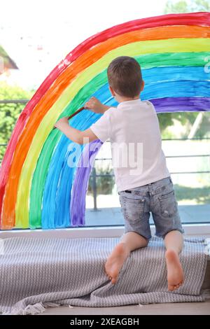 Bambino che disegna l'arcobaleno su una finestra interna, vista posteriore Foto Stock