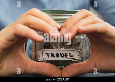 Donna irriconoscibile che mostra un segno cardiaco che risparmia denaro in un vaso di vetro pieno di banconote in dollari. Trascrizione di VIAGGIO DI fronte a. Foto Stock