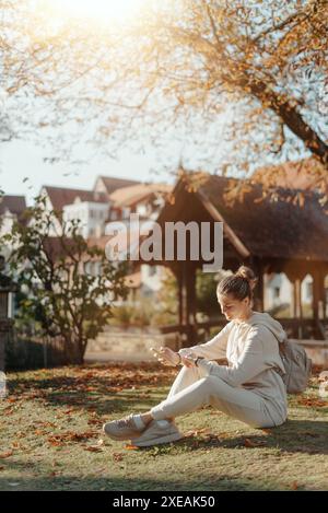 Giovane ragazza adolescente alla moda con smartphone nel Parco Europian in autunno seduta a sorridere. Giovane donna alla moda in autunno a Park Foto Stock