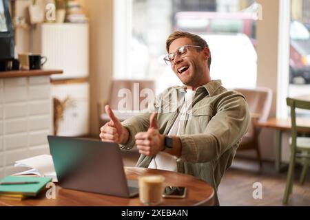 Ritratto di un giovane soddisfatto e felice con gli occhiali, che lavora in un bar, che siede in uno spazio di co-working con un computer portatile, che mostra i pollici in alto, Foto Stock
