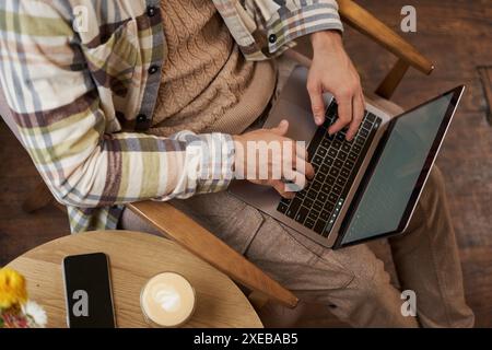 L'angolo superiore ritagliato di mani maschili, l'uomo seduto in un bar con una tazza di caffè e che lavora su un computer portatile, digitando sulla tastiera, ha un Foto Stock
