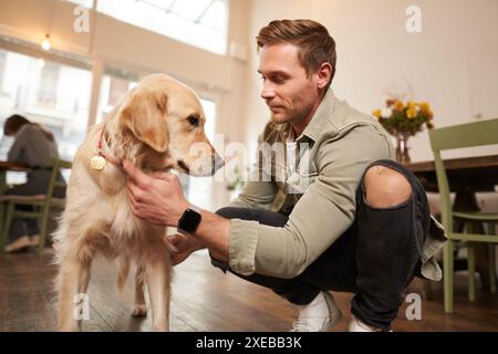 Ritratto di un uomo felice che trascorre del tempo con il suo cane in un bar in cui sono ammessi gli animali domestici, gioca e accarezza Golden retriever, divertiti, regalando Foto Stock