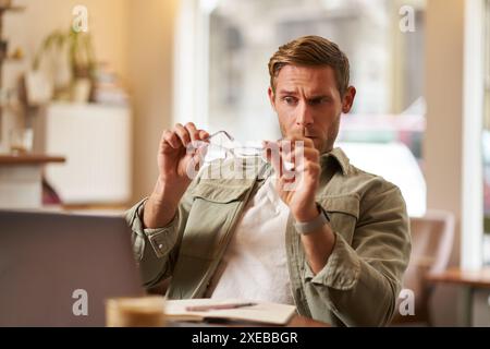 Ritratto di un uomo che sembra confuso attraverso i suoi occhiali, che controlla gli occhiali blu, che siede in un bar con un computer portatile, che lavora su di lui Foto Stock