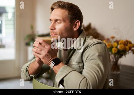 Ritratto di un giovane uomo che chiude gli occhi, puzza di caffè appena fatto in un bar, siede su una sedia con un bicchiere di cappuccino, si gode la t Foto Stock