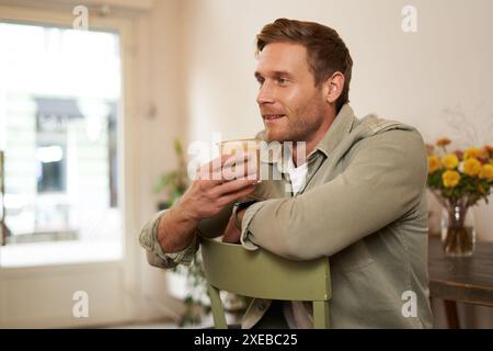 Primo piano ritratto di un bell'uomo, seduto in un bar sulla sedia, sorseggiando un bicchiere di caffè, bevendo un cappuccino e sorridendo. Concetto o Foto Stock