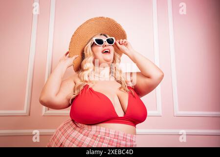 Una donna di taglia Happy Plus indossa abiti estivi in piedi su sfondo rosa mentre tocca gli occhiali da sole e guarda in alto Foto Stock