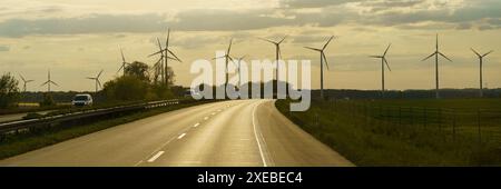 Una strada lunga e rettilinea si snoda attraverso un paesaggio rurale con turbine eoliche in lontananza. Il cielo è nuvoloso e il sole sta tramontando, gettando una g dorata Foto Stock