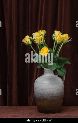 Un bellissimo bouquet di fiori di rose gialle in un vaso grigio su uno sfondo di tessuto marrone scuro. Spazio per testo, copia spazio Foto Stock