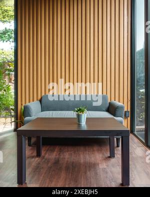 Interni dal design contemporaneo del soggiorno tropicale con divano a cuscino, pianta sul tavolo, parete in legno e giardino tropicale Foto Stock