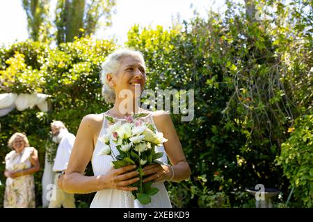 Donna anziana sorridente che tiene un bouquet di fiori alla cerimonia di nozze all'aperto Foto Stock