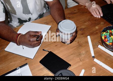 Vista dall'alto dell'irriconoscibile uomo Nero che prende appunti e tiene una tazza di caffè seduto al tavolo di legno nell'area copie per riunioni d'affari Foto Stock