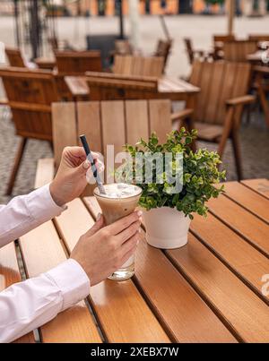Donna che tiene in mano un bicchiere con caffè ghiacciato su un tavolo di legno su una terrazza all'aperto Foto Stock