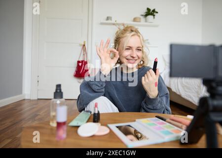 Giovane donna allegra e sorridente che registra un vlog, mostra un gesto ok e il rossetto preferito, consigliando un prodotto di bellezza per Foto Stock