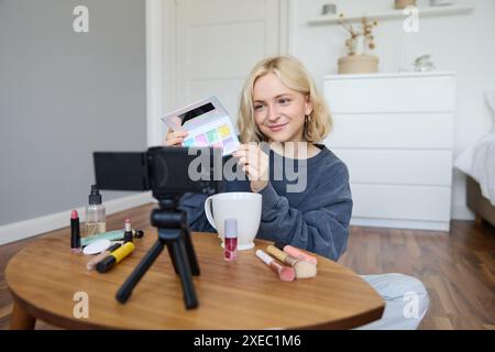 Blogger di lifestyle, registra video nella sua stanza, ha una fotocamera sul tavolino da caffè, mostra la tavolozza delle ombre ai suoi seguaci, fa makeu Foto Stock