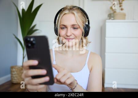 Ritratto ravvicinato di una donna sportiva sorridente che indossa le cuffie, usa il telefono cellulare, app per allenarsi mentre ti alleni a casa Foto Stock