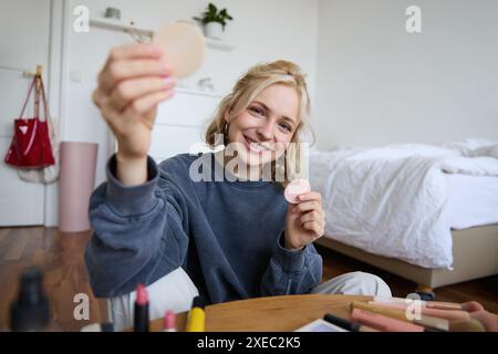 Immagine di una giovane donna bionda, la ragazza registra video sul trucco, mostra prodotti di bellezza per il tono della pelle, siede in una stanza sul pavimento Foto Stock