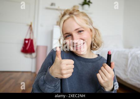 La ragazza sorridente mostra rossetto e pollice in alto, consiglia prodotti di bellezza, registra contenuti per account di social media, Foto Stock