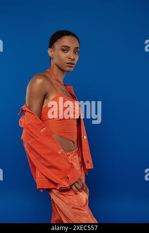 La graziosa giovane donna afroamericana in un vivace abito arancione si mette in posa con sicurezza su uno sfondo blu sorprendente. Foto Stock