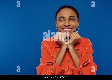 Una giovane donna afroamericana in un vivace vestito arancione sorride, le mani sul viso, irradiando felicità. Foto Stock