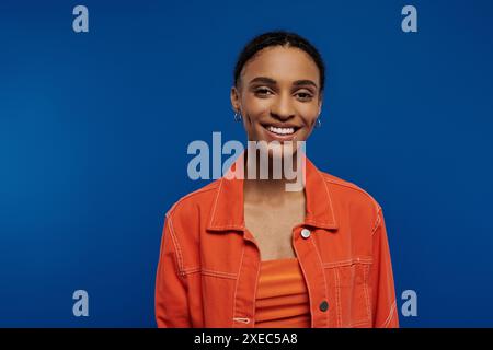 Una giovane donna afroamericana, con una camicia arancione, si proietta felicemente alla telecamera su uno sfondo blu. Foto Stock