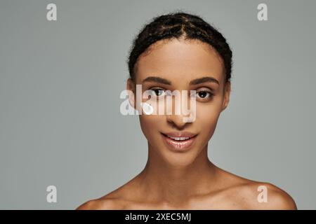 Giovane donna afro-americana con la maglia senza spalline, che le applica delicatamente la crema sul viso su un sereno sfondo grigio. Foto Stock