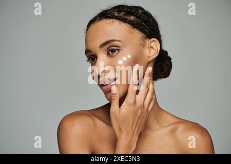 Giovane donna afro-americana con il viso ricoperto di crema, incentrata sulla routine della cura della pelle, in un top senza spalline su sfondo grigio. Foto Stock