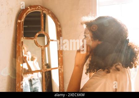 Un primo piano di un'affascinante adolescente mista le mette una maschera cosmetica in tessuto sul viso. Il riflesso di un attraente gir africano Foto Stock