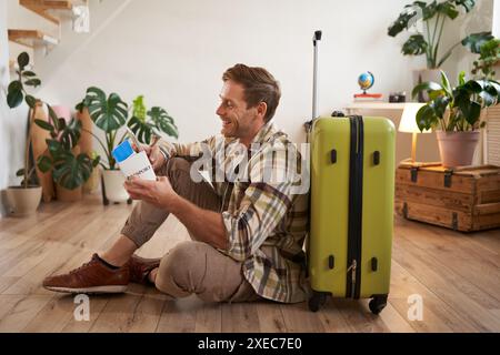 Ritratto di un bell'uomo sorridente, seduto con la valigia sul pavimento, in possesso di passaporto e biglietti aerei, in vacanza Foto Stock