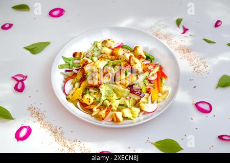 Un'insalata vivace con formaggio halloumi alla griglia e una varietà di verdure fresche, servita in una ciotola bianca con foglie sparse e petali. Foto Stock