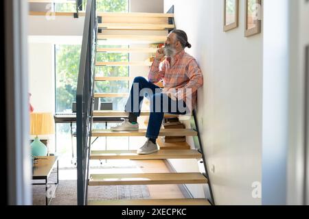 Seduto sulle scale, un anziano beve caffè e guarda fuori dalla finestra di casa Foto Stock