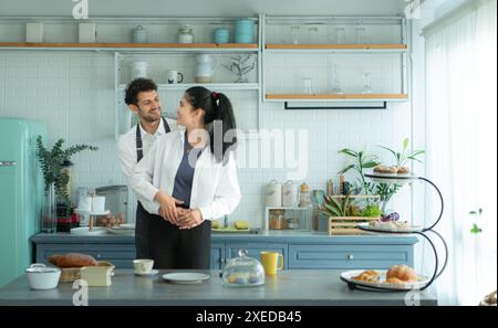 Un marito indossa un grembiule mentre cucina la cena con sua moglie. Ha abbracciato sua moglie felicemente prima di andare in cucina a cucinare. Foto Stock