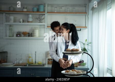 Un marito indossa un grembiule mentre cucina la cena con sua moglie. Ha abbracciato sua moglie felicemente prima di andare in cucina a cucinare. Foto Stock