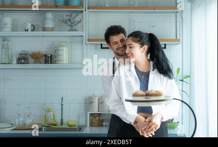 Un marito indossa un grembiule mentre cucina la cena con sua moglie. Ha abbracciato sua moglie felicemente prima di andare in cucina a cucinare. Foto Stock