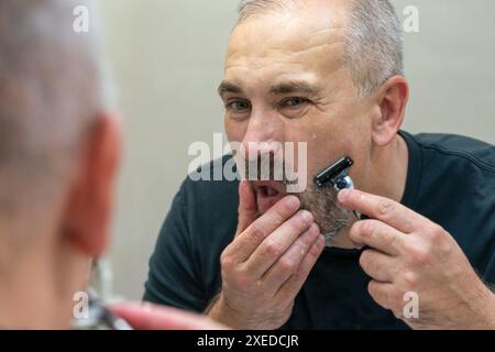 Uomo maturo dai capelli grigi che restyling la sua barba in casa utilizzando il rasoio Foto Stock