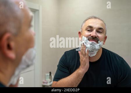 Ritratto di un bell'uomo di mezza età dai capelli grigi che applica schiuma da barba. Affascinante uomo di mezza età che si radica barba e baffi Foto Stock