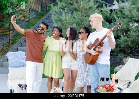 Un gruppo di amici multirazziali che si divertono e bevono vino, scattano selfie durante il picnic estivo. Foto Stock