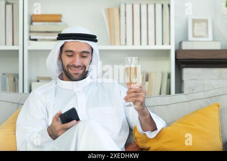 Ritratto di un uomo arabo sorridente che regge un bicchiere di champagne e usa il cellulare a casa Foto Stock