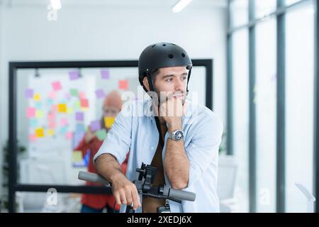 Ritratto di un uomo d'affari sorridente che guida uno scooter elettrico in ufficio con un collega sullo sfondo Foto Stock