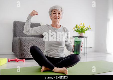 Una donna anziana allegra guarda nella fotocamera e mostra il suo Bicep, incarnando uno stile di vita attivo e gli allenamenti domestici Foto Stock