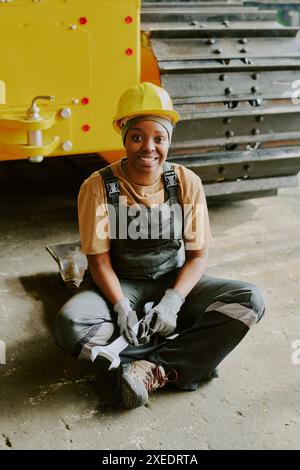 Ripresa verticale di una giovane donna afroamericana felice che indossa un elmetto seduto sul pavimento con la chiave in mano sorridente alla macchina fotografica Foto Stock