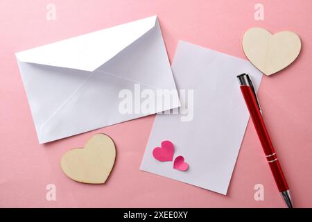 Lo sfondo della lettera sta per scrivere con un foglio bianco vuoto e una busta con penna e ritagli di cuori intorno ad esso su sfondo rosa. Vista dall'alto. Foto Stock