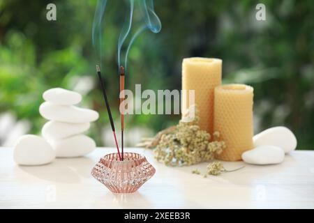 Bastoncini di incenso che si mescolano nel supporto vicino a pietre, candele e fiori secchi su un tavolo di legno all'aperto Foto Stock