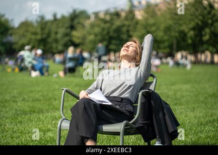 Una donna rilassata riposa su una sedia urbana sul prato si prende una pausa dal lavoro a distanza godendosi il sole a occhi chiusi Foto Stock