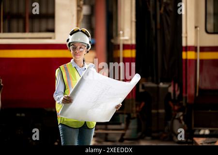 Ritratto del lavoratore tecnico ferroviario in giubbotto di sicurezza e casco che lavora con il progetto alla stazione di riparazione dei treni Foto Stock