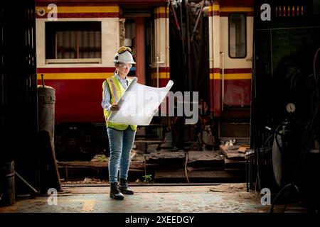 Ritratto del lavoratore tecnico ferroviario in giubbotto di sicurezza e casco che lavora con il progetto alla stazione di riparazione dei treni Foto Stock