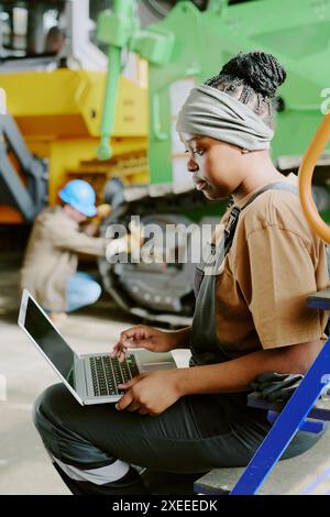Immagine verticale di una giovane donna afroamericana che lavora in una fabbrica moderna controllando i dati su un computer portatile mentre il suo collega ripara la macchina in background Foto Stock
