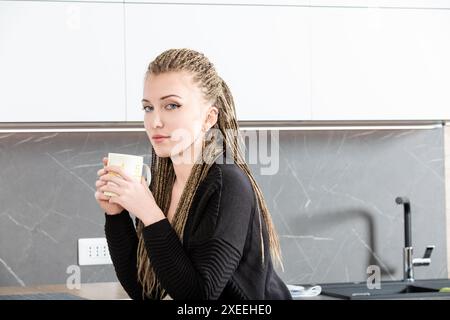 Una giovane donna con trecce bionde si gode una tranquilla pausa caffè nella sua moderna cucina, creando un'atmosfera accogliente e serena Foto Stock